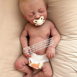 Op de afbeelding: Een realistische babypop met blond haar, gesloten ogen en een speen in de mond. De pop draagt een witte luier en ligt op een beige oppervlak. De afbeelding bevat de tekst "Website: carolineheussdolls.com CarolineHeussDolls | Etsy.com Instagram: @carolineheussdolls Facebook: CarolineHeussDolls"