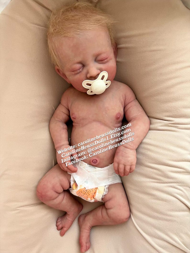Op de afbeelding: Een realistische babypop met blond haar, gesloten ogen en een speen in de mond. De pop draagt een witte luier en ligt op een beige oppervlak. De afbeelding bevat de tekst "Website: carolineheussdolls.com CarolineHeussDolls | Etsy.com Instagram: @carolineheussdolls Facebook: CarolineHeussDolls"