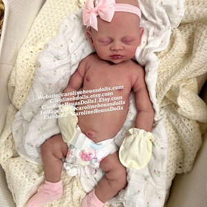 May include: A realistic newborn doll with closed eyes, wearing a light pink headband with a bow, pale yellow mittens, pink socks, and a diaper with a floral pattern. The text on the image reads: Website: carolineheussdolls.com, Carolinehdolls.etsy.com, CarolineHeussDolls1.Etsy.com, Instagram: @carolineheussdolls, Facebook: CarolineHeussDolls.