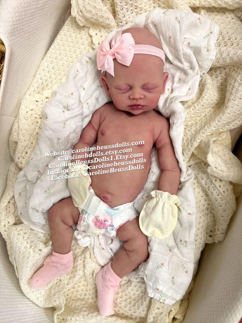 May include: A realistic newborn doll with closed eyes, wearing a light pink headband with a bow, pale yellow mittens, pink socks, and a diaper with a floral pattern. The text on the image reads: Website: carolineheussdolls.com, Carolinehdolls.etsy.com, CarolineHeussDolls1.Etsy.com, Instagram: @carolineheussdolls, Facebook: CarolineHeussDolls.