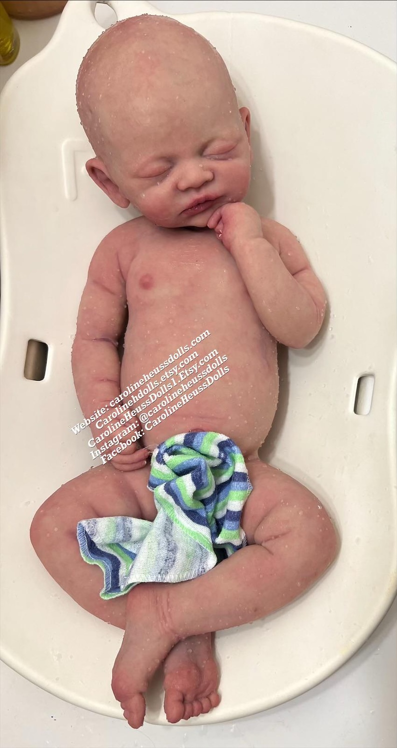 May include: A realistic silicone baby doll with closed eyes, wearing a blue and green striped towel, lying on its back in a white tub.