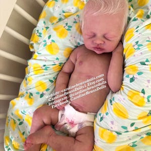 May include: A realistic newborn doll with pale skin and light hair, wearing a white diaper. The doll is lying on a fabric with a yellow lemon and green leaf pattern. Text on the image reads: Website: carolineheussdolls.com, CarolineHeussDolls1.Etsy.com, Instagram: @carolineheussdolls, Facebook: CarolineHeussDolls.