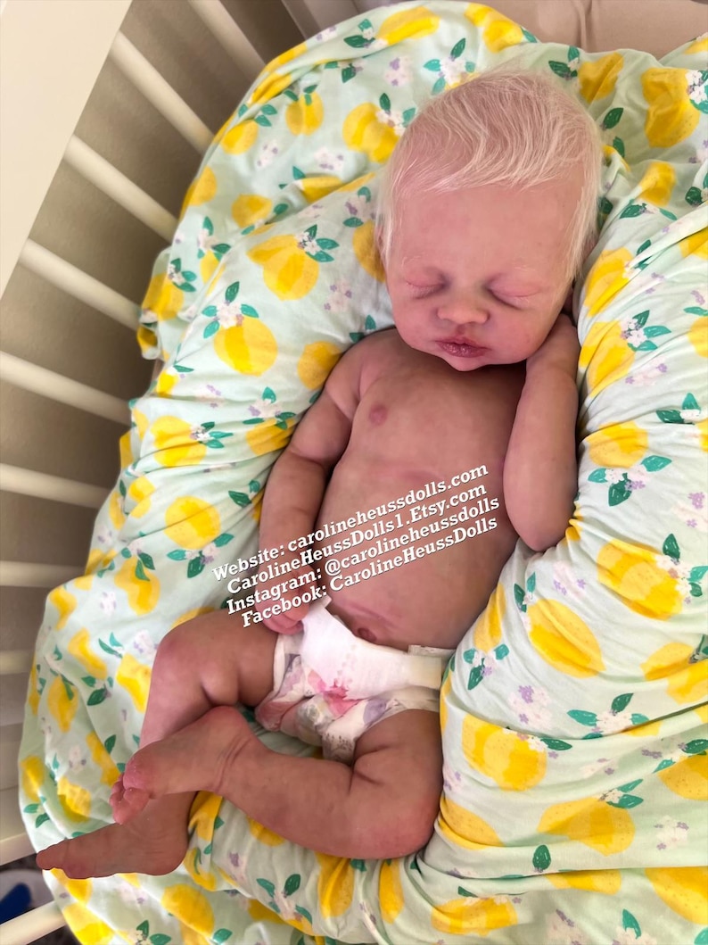 May include: A realistic newborn doll with pale skin and light hair, wearing a white diaper. The doll is lying on a fabric with a yellow lemon and green leaf pattern. Text on the image reads: Website: carolineheussdolls.com, CarolineHeussDolls1.Etsy.com, Instagram: @carolineheussdolls, Facebook: CarolineHeussDolls.
