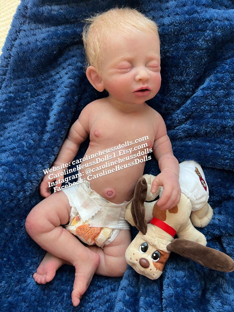 May include: A silicone baby doll with blonde hair wearing a diaper and holding a brown and white plush dog toy. The doll is lying on a blue fuzzy blanket. The text on the blanket reads: 'Website: carolineheussdolls.com CarolineHeussDolls | Etsy.com Instagram: @carolineheussdolls Facebook: CarolineHeussDolls'