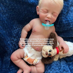 Op de afbeelding: Een realistische reborn babypop met blond haar, die een luier draagt en een pluche hondenspeelgoed vasthoudt. De pop slaapt met een speen in zijn mond. De pop ligt op een blauwe deken. Website: carolineheussdolls.com CarolineHeussDolls1.Etsy.com Instagram: @carolineheussdolls Facebook: CarolineHeussDolls