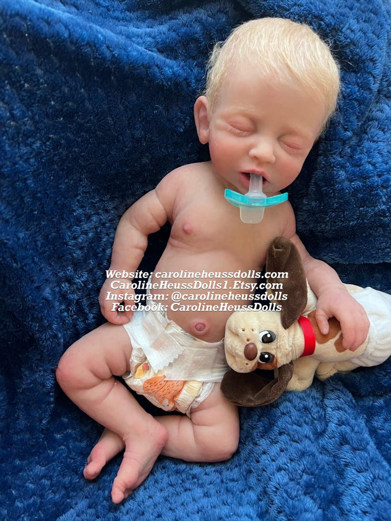 Op de afbeelding: Een realistische reborn babypop met blond haar, die een luier draagt en een pluche hondenspeelgoed vasthoudt. De pop slaapt met een speen in zijn mond. De pop ligt op een blauwe deken. Website: carolineheussdolls.com CarolineHeussDolls1.Etsy.com Instagram: @carolineheussdolls Facebook: CarolineHeussDolls