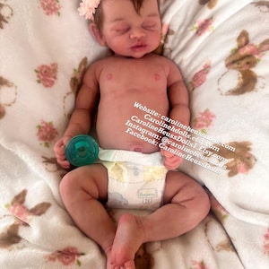 May include: A realistic newborn baby doll with light brown hair, wearing a pale pink floral headband and a white diaper. The doll holds a small teal pacifier in one hand. Text on the image reads: Website: carolineheussdolls.com, Carolineheussdolls.Etsy.com, CarolineHeussDolls1.Etsy.com, Instagram: @carolineheussdolls, Facebook: CarolineHeussDolls.
