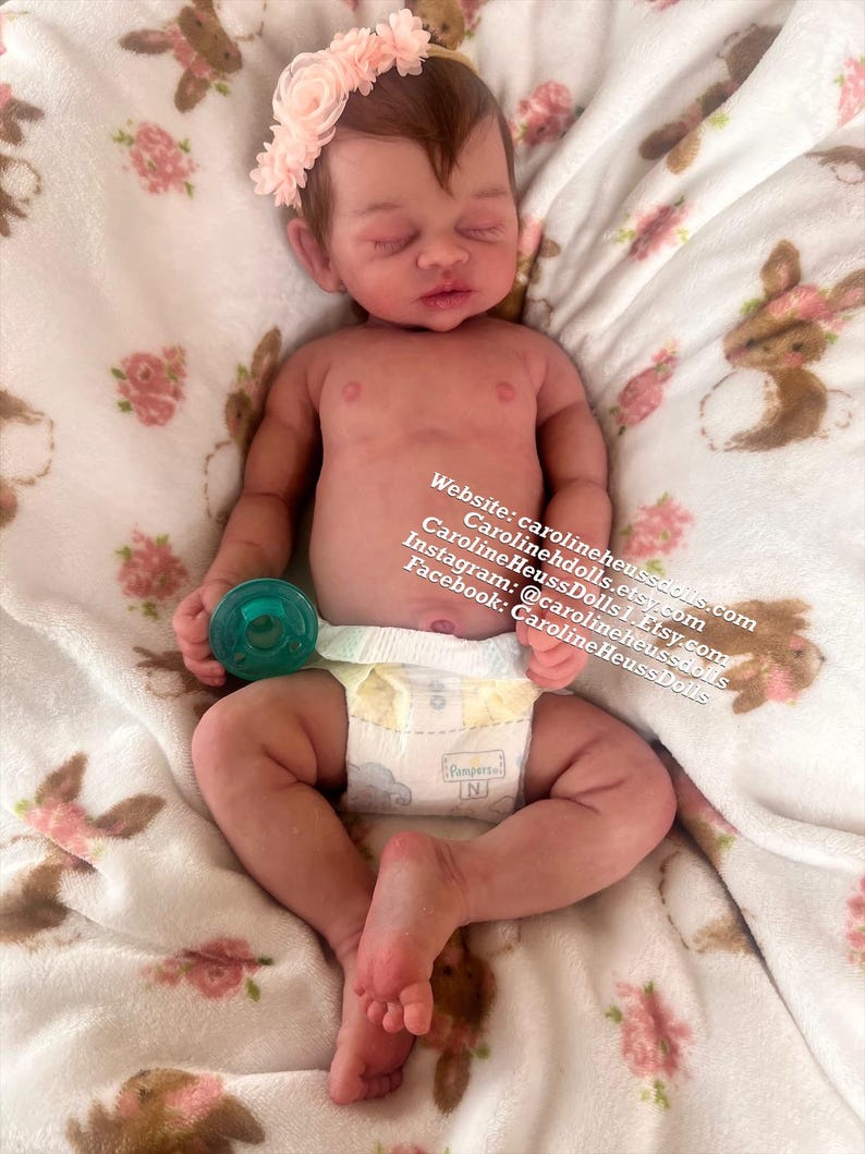 May include: A realistic newborn baby doll with light brown hair, wearing a pale pink floral headband and a white diaper. The doll holds a small teal pacifier in one hand. Text on the image reads: Website: carolineheussdolls.com, Carolineheussdolls.Etsy.com, CarolineHeussDolls1.Etsy.com, Instagram: @carolineheussdolls, Facebook: CarolineHeussDolls.