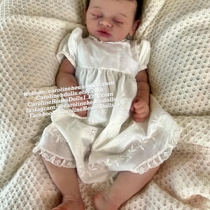 May include: A realistic newborn doll with closed eyes and dark hair, wearing a white short-sleeved dress with eyelet embroidery and lace trim. Text on the image reads: Website: carolineheussdolls.com, Carolinehdolls.etsy.com, CarolineHeussDolls1.Etsy.com.