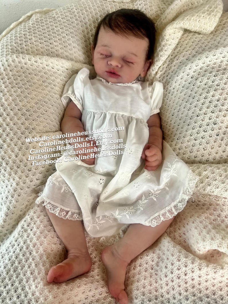 May include: A realistic newborn doll with closed eyes and dark hair, wearing a white short-sleeved dress with eyelet embroidery and lace trim. Text on the image reads: Website: carolineheussdolls.com, Carolinehdolls.etsy.com, CarolineHeussDolls1.Etsy.com.