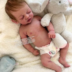 May include: A realistic newborn doll with light skin, reddish-brown hair, and closed eyes, wearing a patterned diaper. The doll is positioned next to a plush gray rabbit toy. Text on the doll reads: Website: carolineheussdolls.com, Carolineheussdolls.etsy.com, CarolineHeussDolls1.Etsy.com, Instagram: @carolineheussdolls, Facebook: CarolineHeussDolls.