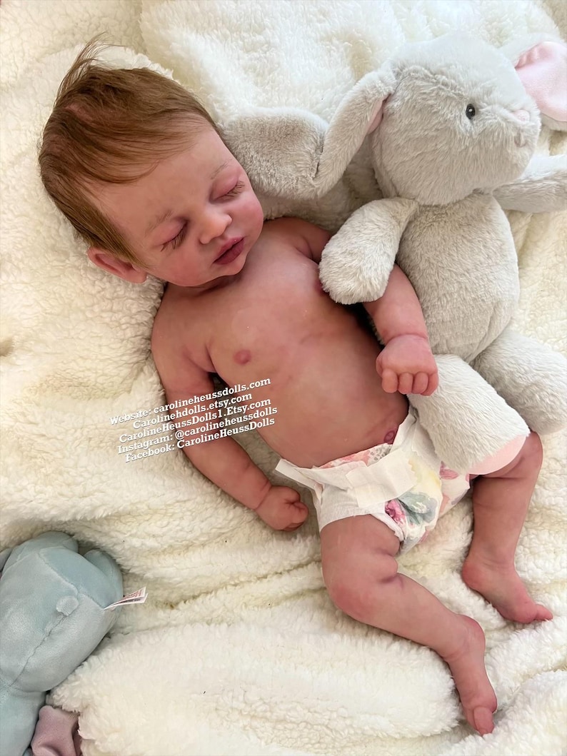 May include: A realistic newborn doll with light skin, reddish-brown hair, and closed eyes, wearing a patterned diaper. The doll is positioned next to a plush gray rabbit toy. Text on the doll reads: Website: carolineheussdolls.com, Carolineheussdolls.etsy.com, CarolineHeussDolls1.Etsy.com, Instagram: @carolineheussdolls, Facebook: CarolineHeussDolls.