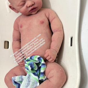 May include: A realistic newborn baby doll with pale skin, closed eyes, and subtle mottling, lying on a white plastic bath seat. A small blue, green, and white striped cloth covers the lower body. Text includes carolineheussdolls.com and social media handles.