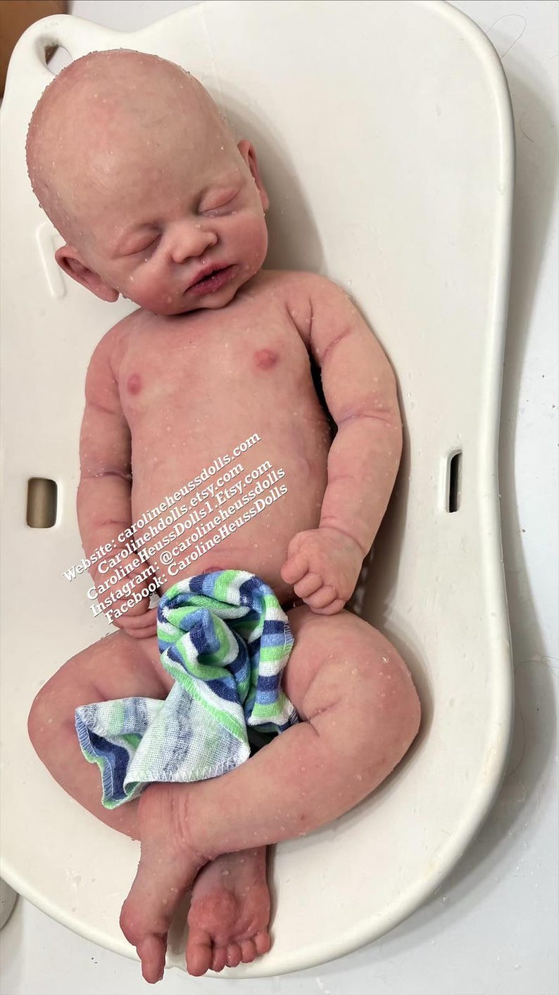 May include: A realistic newborn baby doll with pale skin, closed eyes, and subtle mottling, lying on a white plastic bath seat. A small blue, green, and white striped cloth covers the lower body. Text includes carolineheussdolls.com and social media handles.