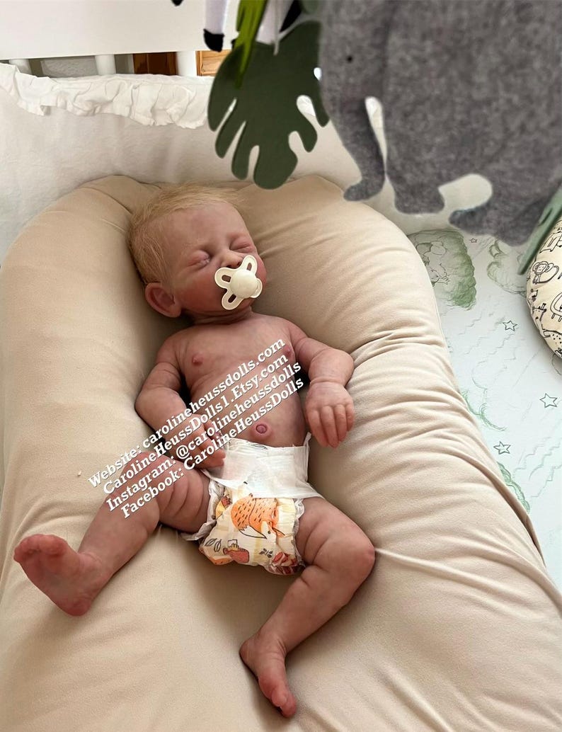 May include: A realistic baby doll with closed eyes and a pacifier in its mouth. The doll is wearing a diaper with a fox print and is lying on a tan pillow. The text on the image reads: 'Website: carolineheussdolls.com CarolineHeussDolls | Etsy.com Instagram: @carolineheussdolls Facebook: CarolineHeussDolls'