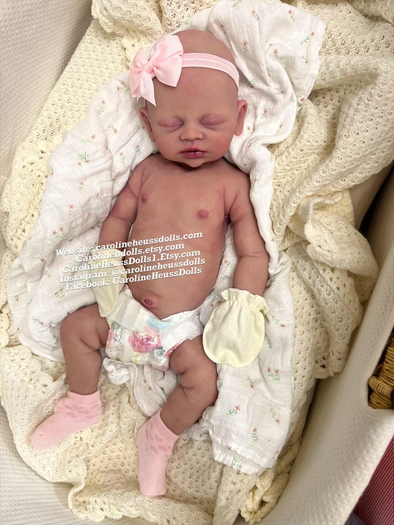 May include: A realistic newborn baby doll with closed eyes, wearing a light pink headband with a bow, pale yellow mittens, pink socks, and a diaper with a floral pattern. The text on the image reads: Website: carolineheussdolls.com, CarolineHeussDolls.Etsy.com, Instagram: @carolineheussdolls, Facebook: CarolineHeussDolls.