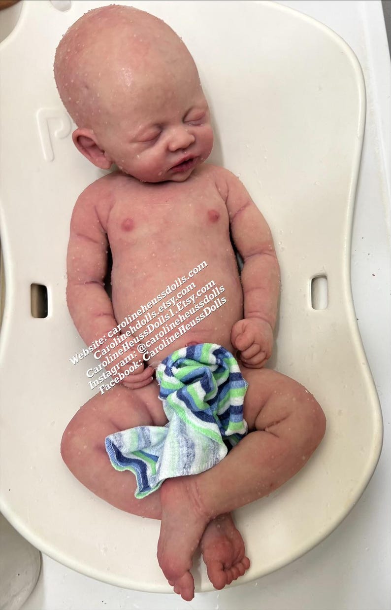 May include: A realistic newborn baby doll with pale skin, closed eyes, and subtle mottling, lying on a white plastic surface. A small blue, green, and white striped cloth covers the lower body. Text on the doll reads: Website: carolineheussdolls.com, carolinehdolls.etsy.com, CarolineHeussDolls1.Etsy.com, Instagram: @carolineheussdolls, Facebook: CarolineHeussDolls.