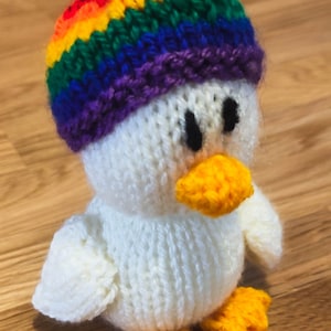 May include: A white knitted bird wearing a rainbow knit hat. The bird has a yellow beak and feet.