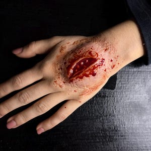 Op de afbeelding: Een hand met een diepe, open wond, realistisch weergegeven met rode en roze tinten. De wond is gecentreerd op de handpalm, met gesimuleerd bloed en blootgesteld weefsel. De huid rond de wond is licht verkleurd.