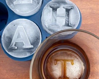 May include: A blue silicone ice cube tray with four ice cubes, each with a letter of the word "LATH" frozen inside. A glass with a brown liquid and a single ice cube with the letter "T" frozen inside.