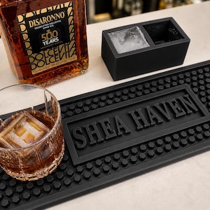 May include: A bar scene with a glass of amber drink and a large ice cube, a bottle of Disaronno liqueur, a black ice cube tray, and a black bar mat with the text "SHEA HAVEN".