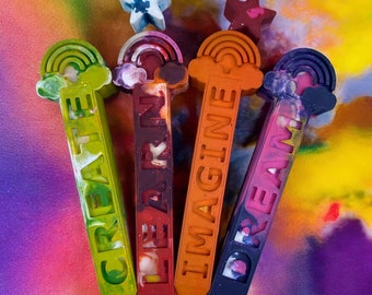 May include: Four colorful crayon sticks with rainbow designs and the words "CREATE", "LEARN", "IMAGINE", and "DREAM" written on them.