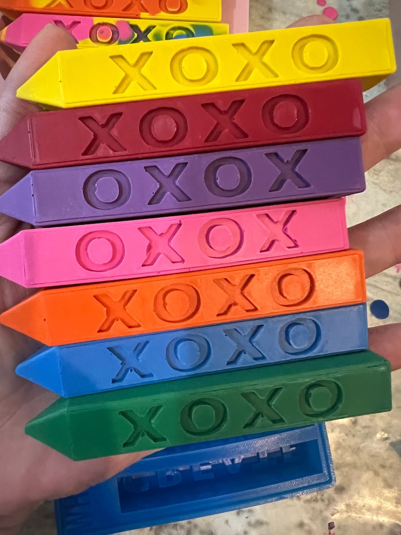 Pu&ograve; includere: Un set di sette pastelli, ognuno con la parola "XOXO" in rilievo. I pastelli sono disposti in ordine arcobaleno, iniziando dal giallo e terminando con il verde.