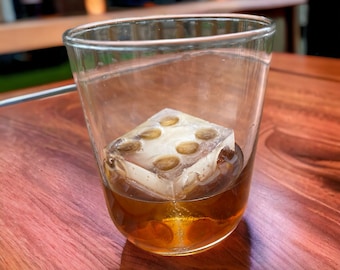 May include: A glass with a brown liquid and a large ice cube with a unique design. The ice cube has a square shape with rounded edges and six circular indentations.