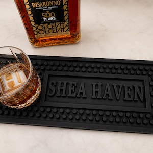 May include: A black bar mat with the text "SHEA HAVEN" is displayed. A glass with a large ice cube and a bottle of Disaronno liqueur are also visible. The liqueur bottle has text that reads "500 YEARS" and "DOLCEVITA".