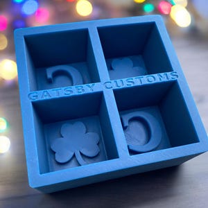 May include: A blue square ice cube tray with four compartments. Each compartment features a different design: a letter "C", a shamrock, and the text "Gatsby Customs". The tray is made of plastic.