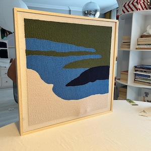 Può includere: Un'opera d'arte da parete quadrata con un design a punch needle testurizzato in una cornice di legno chiaro. La composizione astratta mostra forme stratificate in verde oliva, blu medio, blu navy scuro e crema, creando un motivo paesaggistico.