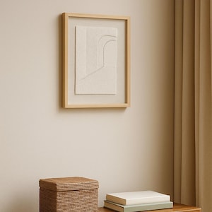 May include: A framed abstract art piece with a textured, off-white design hangs on a beige wall. Below, a woven box with a lid and two books rest on a wooden table. The scene is bathed in soft, natural light.