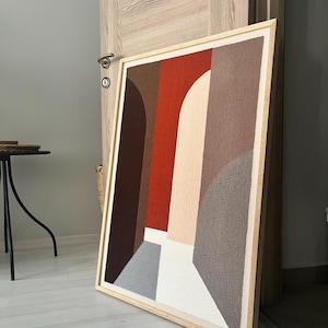 May include: Abstract framed artwork featuring geometric shapes in shades of brown, red, gray, and white. The artwork is displayed in a light wooden frame and leans against a wall. The artwork's design includes arches and rectangular shapes.