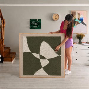 May include: A square wall art piece featuring a textured, tufted design with abstract, overlapping shapes in dark olive green and cream. The artwork is encased in a light-toned wooden frame, showcasing a tactile, rug-like surface with clean lines.