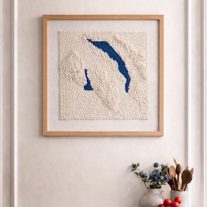 Könnte beinhalten: Gerahmtes, strukturiertes Wandkunstwerk mit hellem Holzrahmen. Das Kunstwerk zeigt einen cremefarbenen Hintergrund mit einer blauen, abstrakten Form. Dekorative Gegenstände, darunter eine Vase mit blauen Blumen und Holzlöffel, befinden sich auf einer dunklen Holzoberfläche.