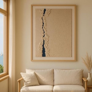 May include: A framed abstract wall art piece with a beige textured surface and a dark blue vertical line. The artwork is in a light wooden frame and hangs above a light-colored sofa. The room has a neutral color palette with natural light coming from a window.
