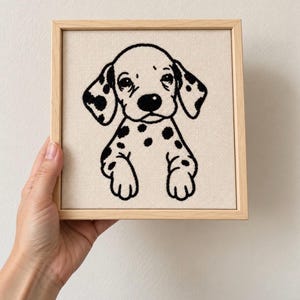 May include: A square wooden frame containing a piece of cream-colored fabric with a black embroidered design of a spotted puppy. The puppy is depicted from the chest up with its front paws resting at the bottom edge of the embroidered area.
