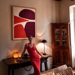May include: A red dress with a scoop neckline. The dress is worn in a room with a large abstract art piece with red and purple shapes. A wooden table with a lamp and candles is in the foreground. A wooden cabinet is in the background.