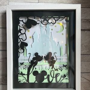 May include: A white frame with a black silhouette of Mickey and Minnie Mouse in front of a castle with a moon and stars. The word "love" is written in a stylized font below the couple.