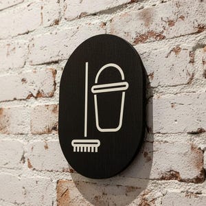 Puede incluir: Un letrero negro de forma ovalada que presenta un icono de líneas blancas de un cubo y una escoba. El letrero tiene un acabado mate suave y está montado sobre una pared de ladrillo blanco texturizado, mostrando una representación gráfica clara.
