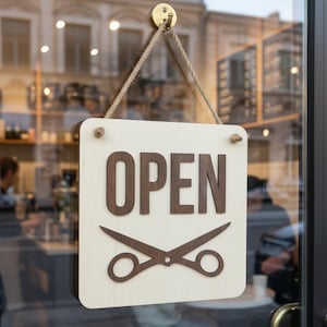 Peut inclure: Panneau "OUVERT" en bois avec lettrage marron et ciseaux, suspendu à une porte vitrée. Le panneau est de couleur crème et suspendu par une corde. L'arrière-plan montre un intérieur flou avec des personnes.