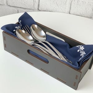May include: A dark brown wooden cutlery holder with a navy blue napkin and silverware. The holder has a rectangular shape with cut-out handles. The napkin has a white floral design and holds two spoons and a fork.