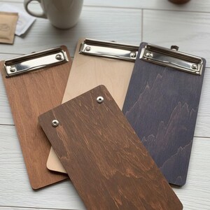 May include: A set of four rectangular wooden clipboards featuring varying wood grain finishes in light, medium, and dark brown tones. Each clipboard is equipped with a silver-colored metal clip at the top, and one board features two silver rivets.