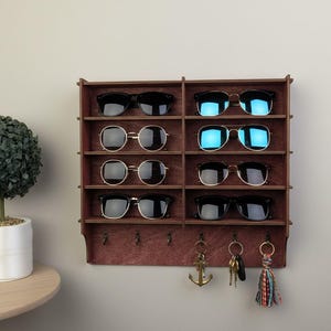May include: A dark brown wooden wall-mounted organizer featuring eight horizontal shelves for storing sunglasses and five metal hooks at the bottom for hanging keys. The shelves currently hold eight pairs of sunglasses with various frame styles.