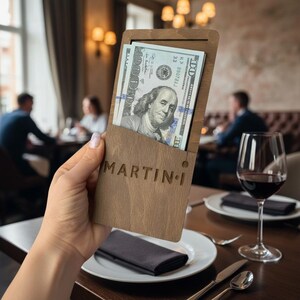 May include: A rectangular wooden bill presenter featuring a laser-engraved logo that reads MARTIN-i. The holder contains a stack of United States one hundred dollar bills, with the portrait of Benjamin Franklin clearly visible at the top of the stack.