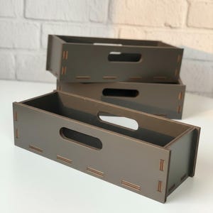 May include: Three brown wooden storage boxes with rectangular cut-out handles. The boxes are stacked, with one in the foreground and two behind it. The boxes are made of wood and have a simple, modern design.