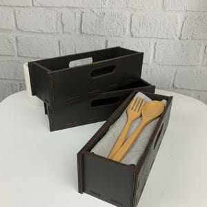 May include: Three dark brown wooden rectangular storage boxes. One box contains a wooden fork and spoon on a gray cloth. The boxes have handle cutouts. The boxes are on a white surface.
