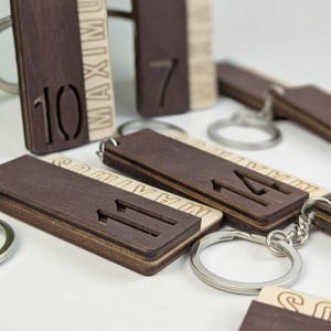 Peut inclure: Une collection de porte-clés rectangulaires en bois composés d'une couche de bois brun foncé sur du bois clair. Chaque porte-clés présente un numéro découpé au laser et le mot MAXIMUM gravé verticalement. Un anneau en métal argenté est fixé.
