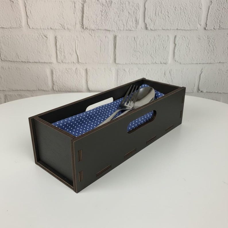 May include: A dark brown wooden cutlery holder with a rectangular shape and cut-out handles. Inside, a blue cloth with white polka dots holds a fork and spoon. The holder sits on a white surface against a white brick wall.