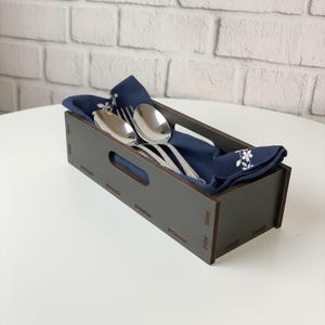 May include: A dark gray wooden cutlery holder with a rectangular shape and cut-out handles. Inside, a folded navy blue napkin with a white floral pattern holds a spoon, fork, and knife. The holder sits on a white surface.